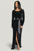 Front view of model wearing black Julia sequin velvet maxi dress with round neckline, long sleeves, elastic waist and right-side slit.