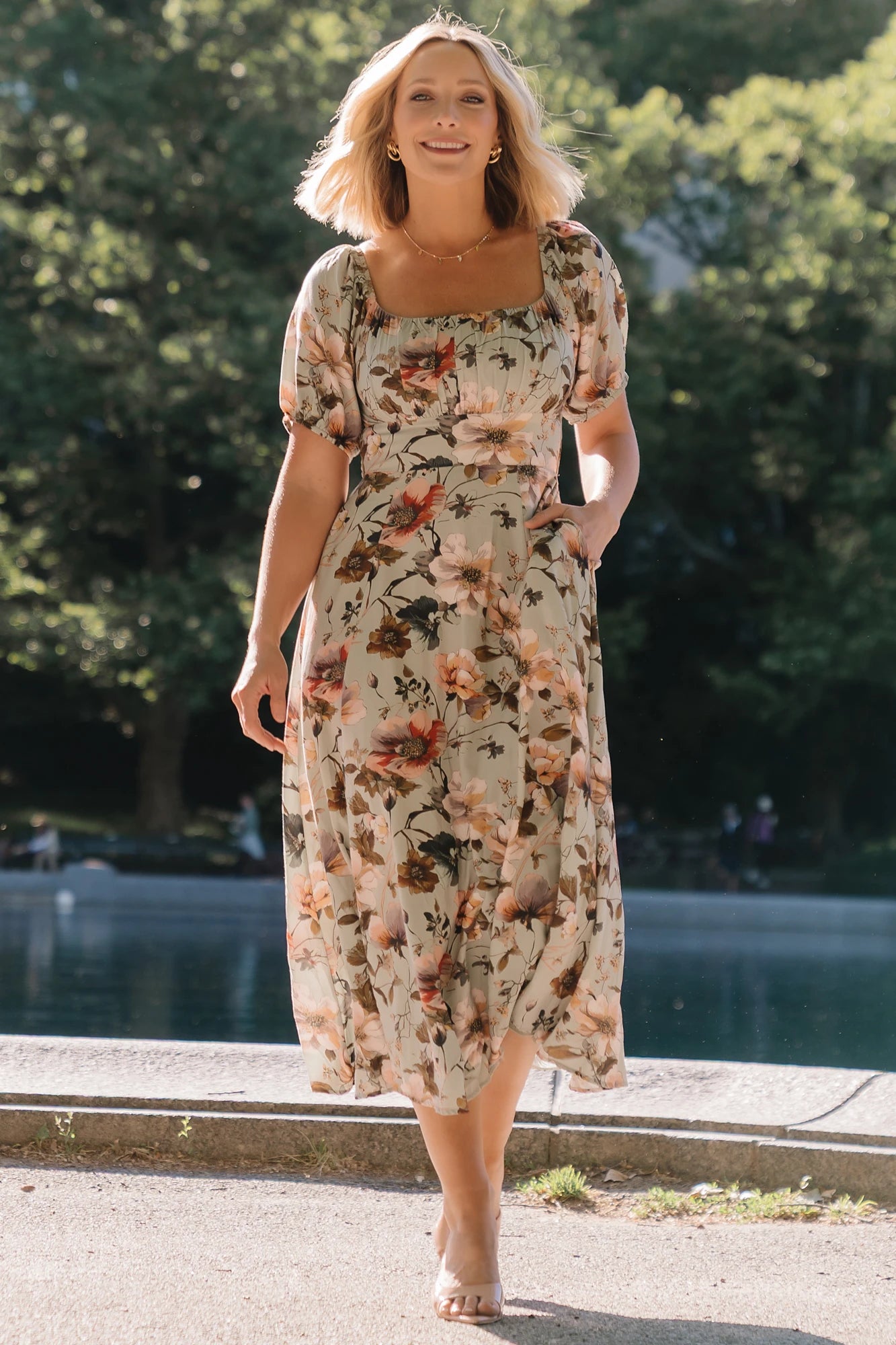 Model wearing the Charlotte Midi Dress in Sage, a sage green midi dress with a pink multi floral print, rounded square neckline, puff sleeves, banded waist, and flowing A-line skirt with side pockets.
