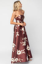 Model wearing a strapless maxi gown with a mahogany floral print and a fitted bodice. A rosette detail accents the neckline, while the flowing floor-length skirt creates elegant movement.