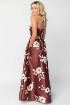 Model wearing a strapless maxi gown with a mahogany floral print and a fitted bodice. A rosette detail accents the neckline, while the flowing floor-length skirt creates elegant movement.