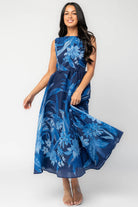 Model wearing a navy sleeveless midi dress with a high boat neckline and a fitted bodice. A tonal blue floral print adds subtle dimension throughout, while the defined waist flows into a full midi skirt that creates graceful movement and an elegant, timeless silhouette.