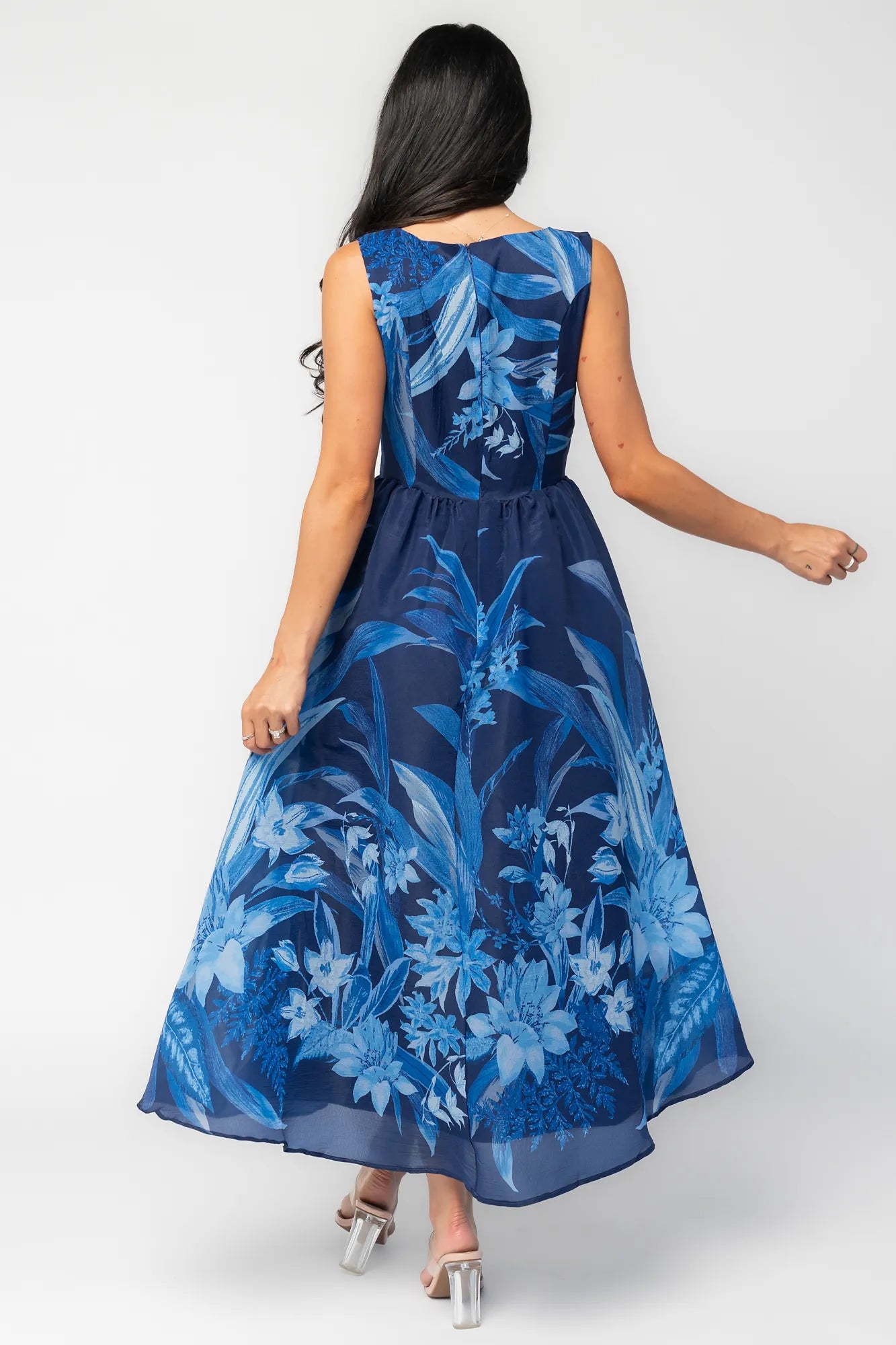 Model wearing a navy sleeveless midi dress with a high boat neckline and a fitted bodice. A tonal blue floral print adds subtle dimension throughout, while the defined waist flows into a full midi skirt that creates graceful movement and an elegant, timeless silhouette.