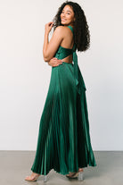 Model walking in emerald pleated maxi dress to show movement of the unlined pleated skirt, drape, and full maxi length in motion.