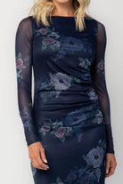 Model wearing a deep plum floral maxi dress with a fitted bodycon silhouette, sheer long sleeves, and a round neckline.