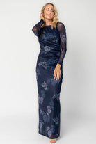 Model wearing a deep plum floral maxi dress with a fitted bodycon silhouette, sheer long sleeves, and a round neckline.