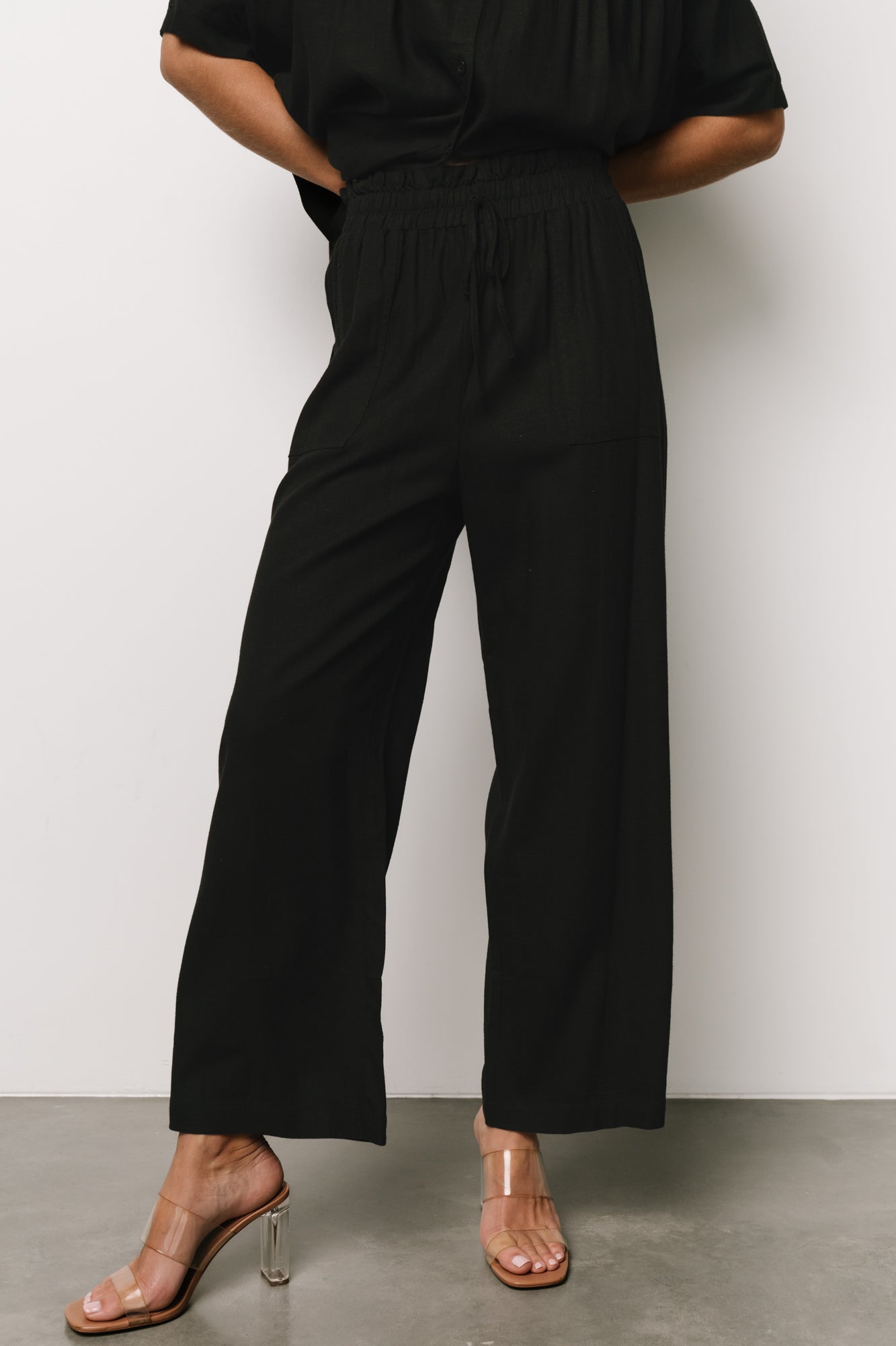 Front full-length view of Romy wide-leg black pants showing smocked elastic waist, faux tie, and front pockets in natural light.
