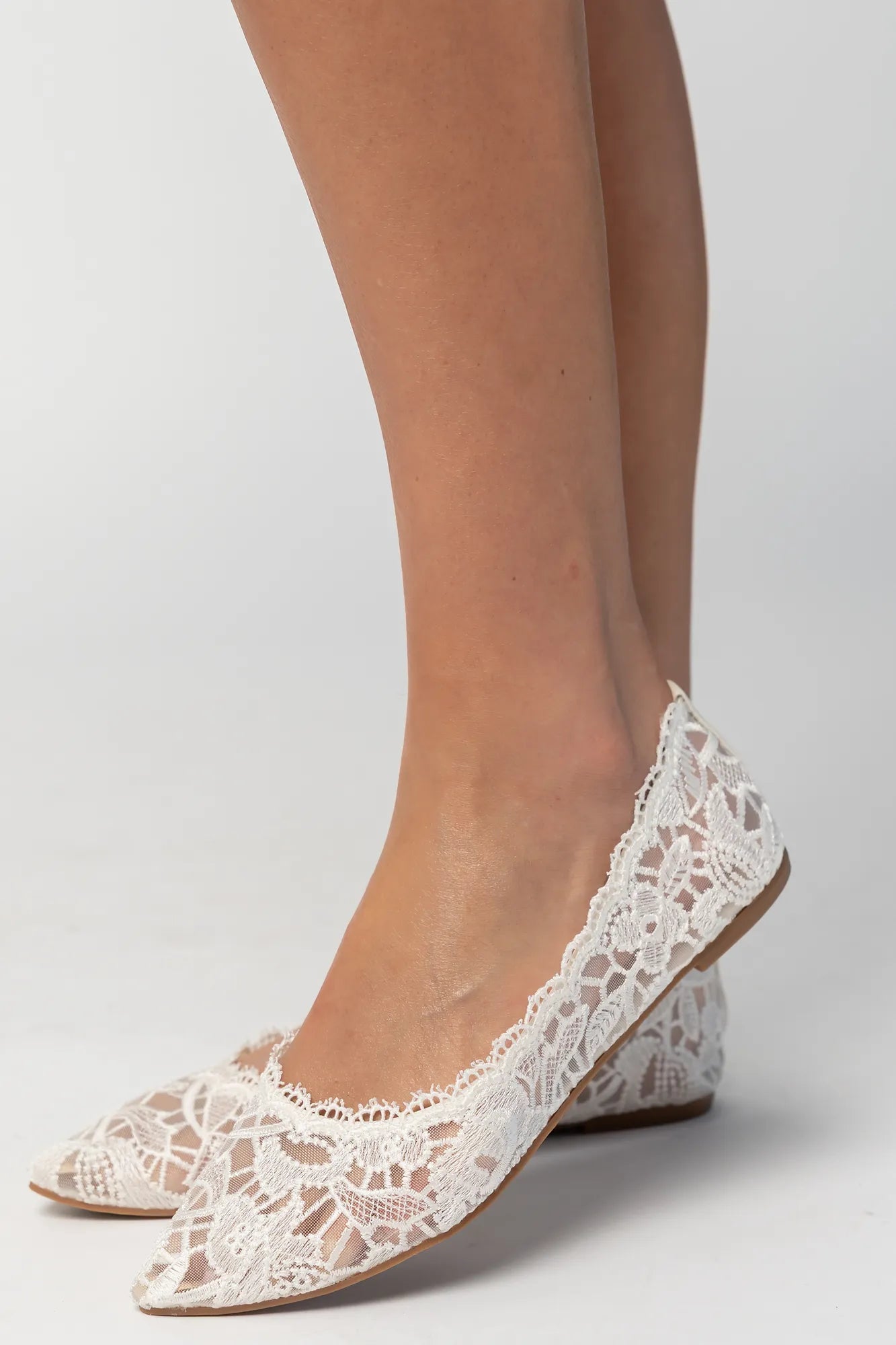 Model wearing ivory lace ballet flats with a pointed toe, sheer lace construction, and scalloped edges. The feminine flat silhouette offers an elegant, lightweight look.