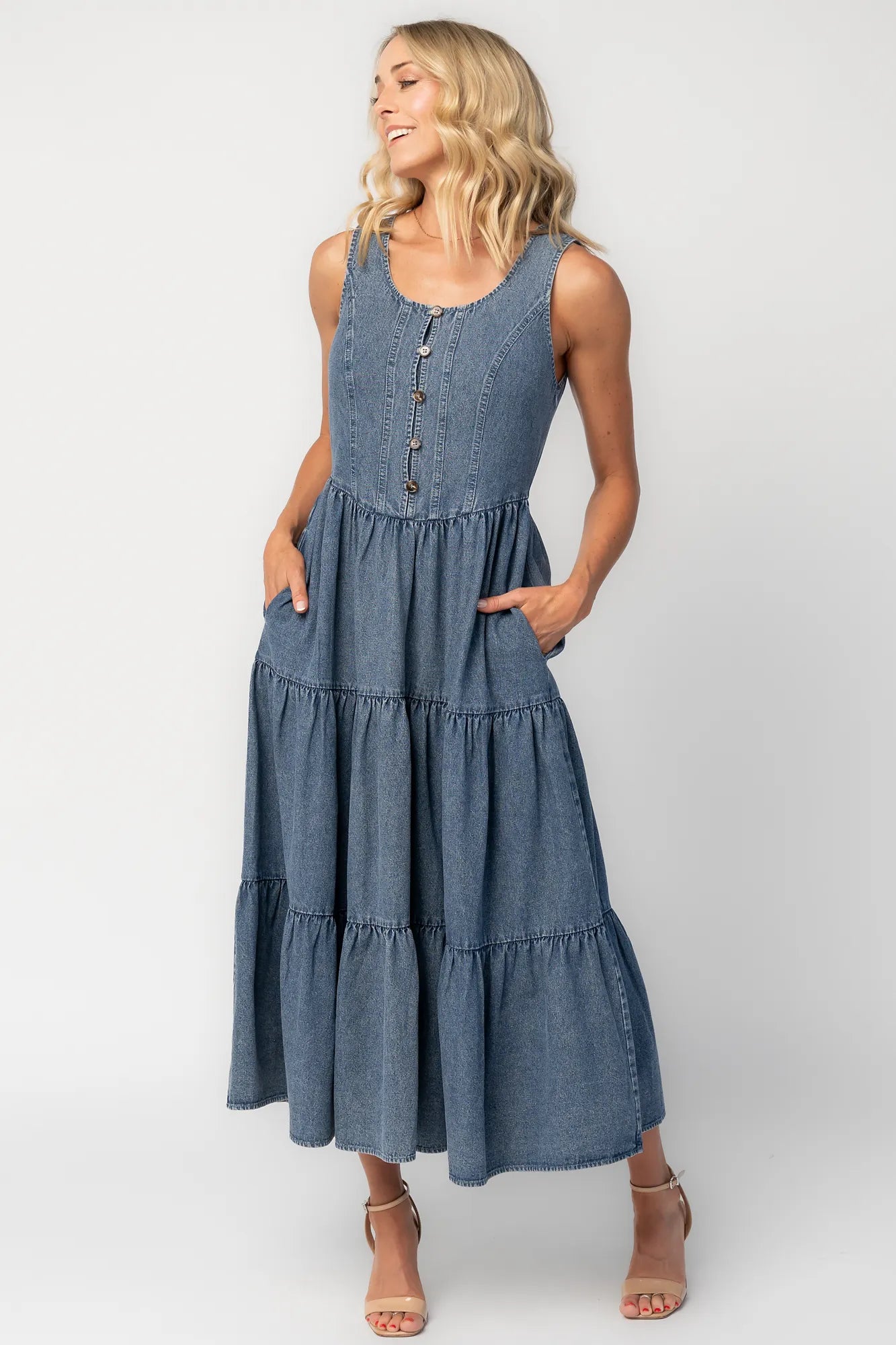 Woman wearing a sleeveless blue denim midi dress with a round neckline, button-front bodice, side pockets, and a tiered skirt.
