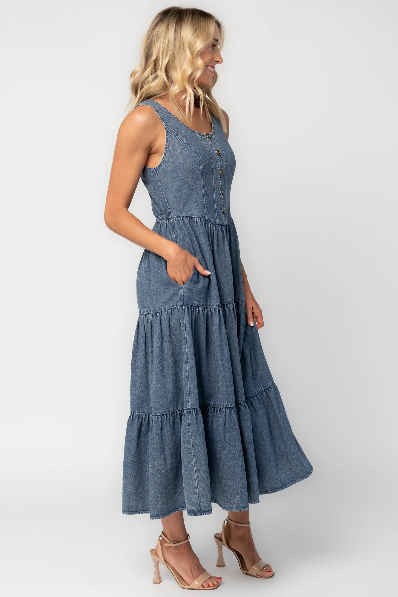 Side image of a woman wearing a sleeveless blue denim midi dress with a round neckline, button-front bodice, side pockets, and a tiered skirt.