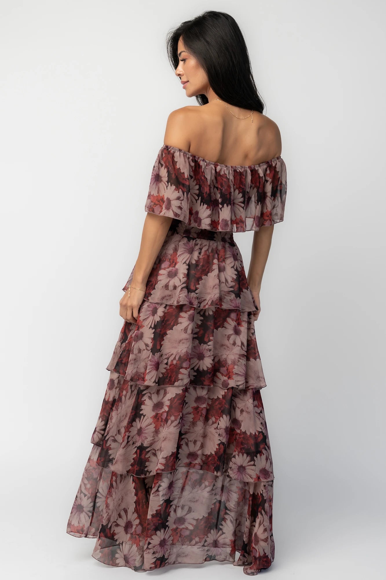 Model wearing the Carina Off-Shoulder Maxi in a soft multi floral print featuring a versatile neckline that can be worn on or off the shoulders, a ruffle overlay across the bust, a smocked waist, and a flowing four-tier maxi skirt.