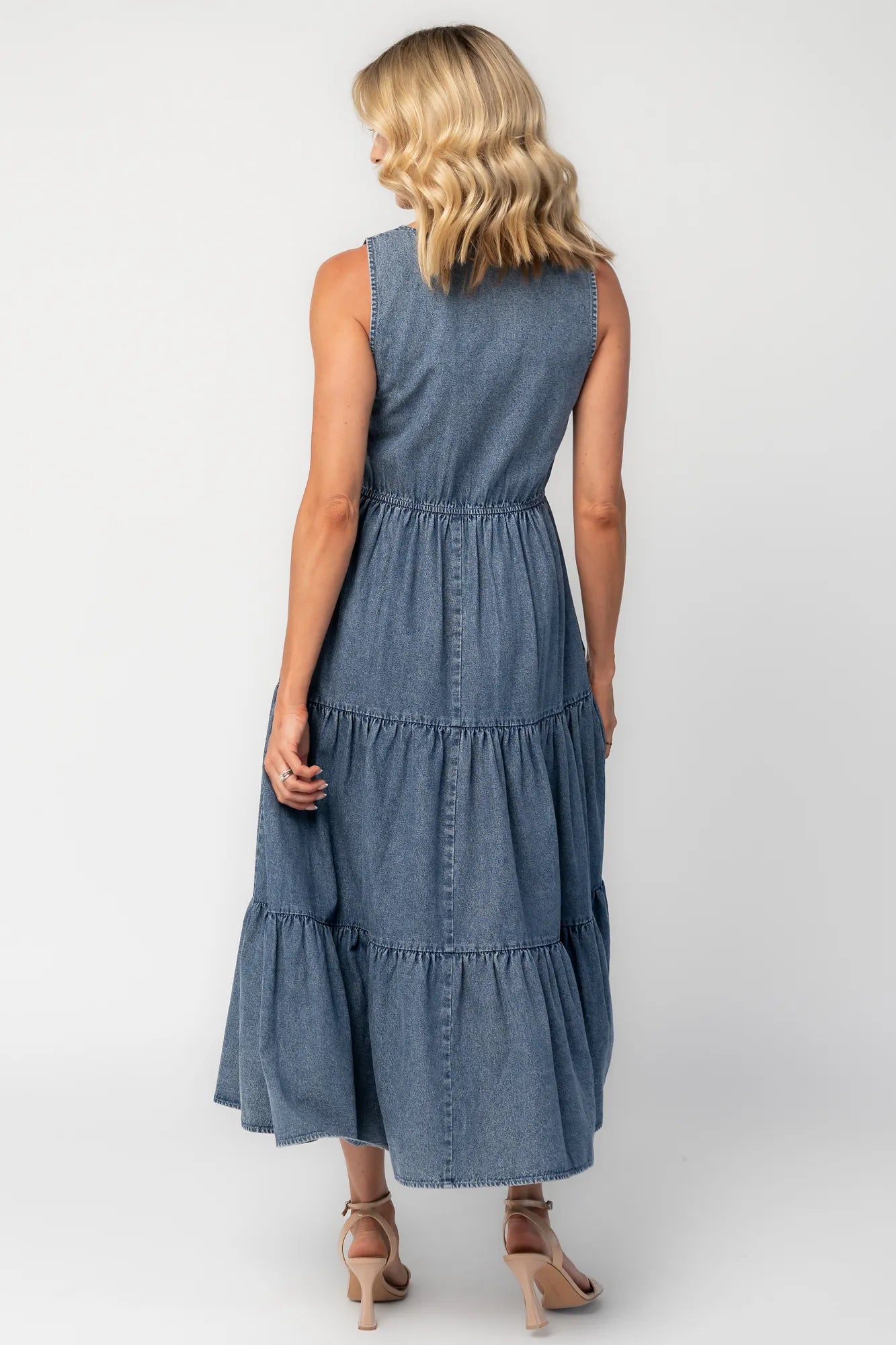 Back image of a woman wearing a sleeveless blue denim midi dress with a round neckline, button-front bodice, side pockets, and a tiered skirt.