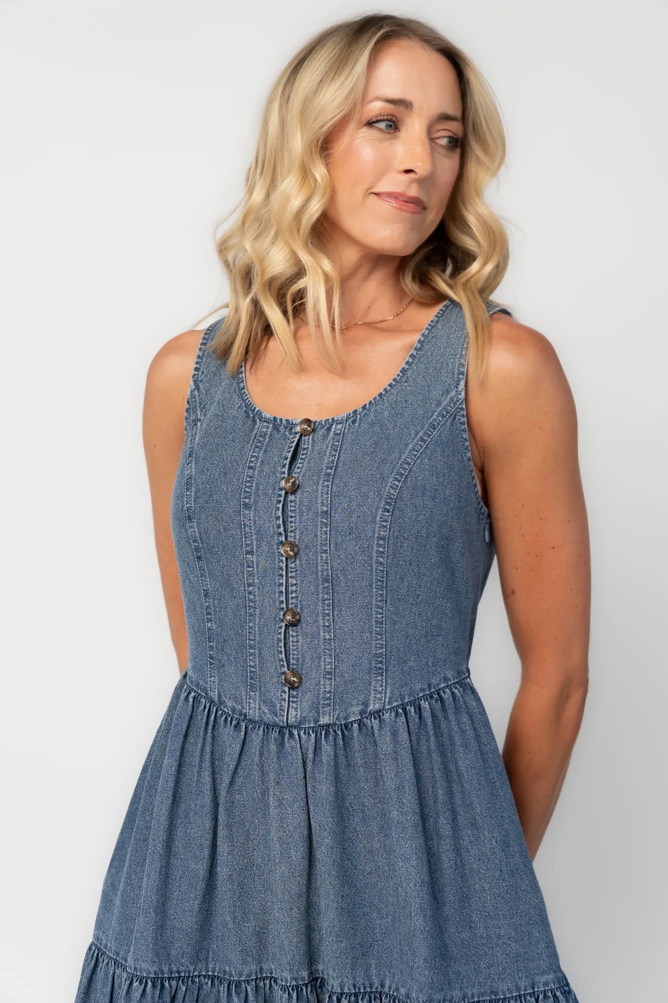 Cropped image of woman wearing a sleeveless blue denim midi dress with a round neckline, button-front bodice, side pockets, and a tiered skirt.