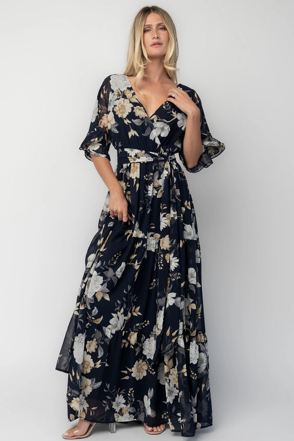 Model wearing the Luna Maxi Dress in Navy Floral featuring a surplice V-neckline with a V-back and tie detail, soft flutter sleeves, and an elastic waist with a removable sash tie