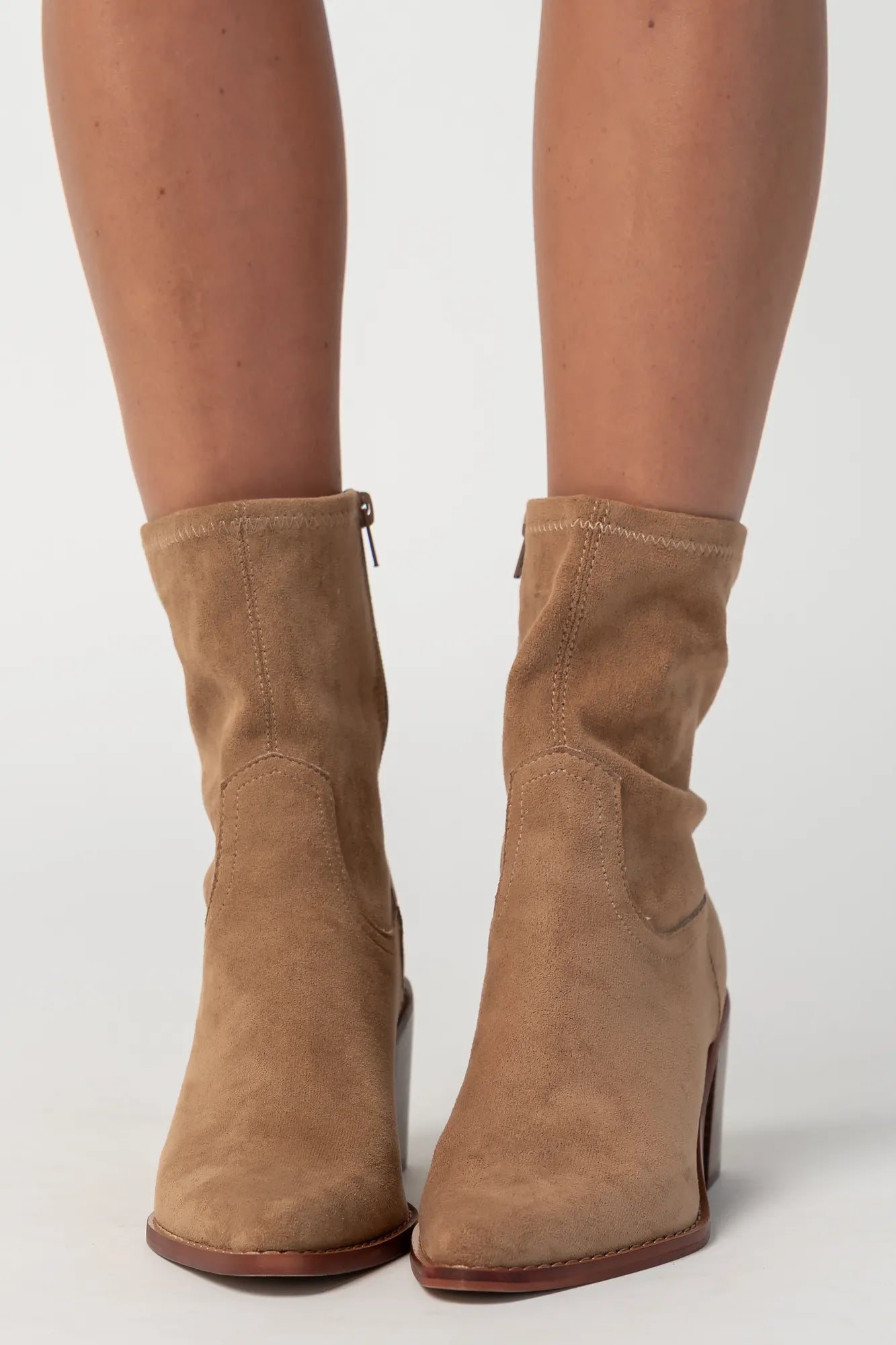 Pair of sand-colored ankle boots featuring a soft suede-like finish, pointed toe silhouette, and stacked block heel. The boots include a side zipper closure and subtle western-inspired seam detailing, creating a versatile look that pairs easily with denim, dresses, and seasonal outfits.