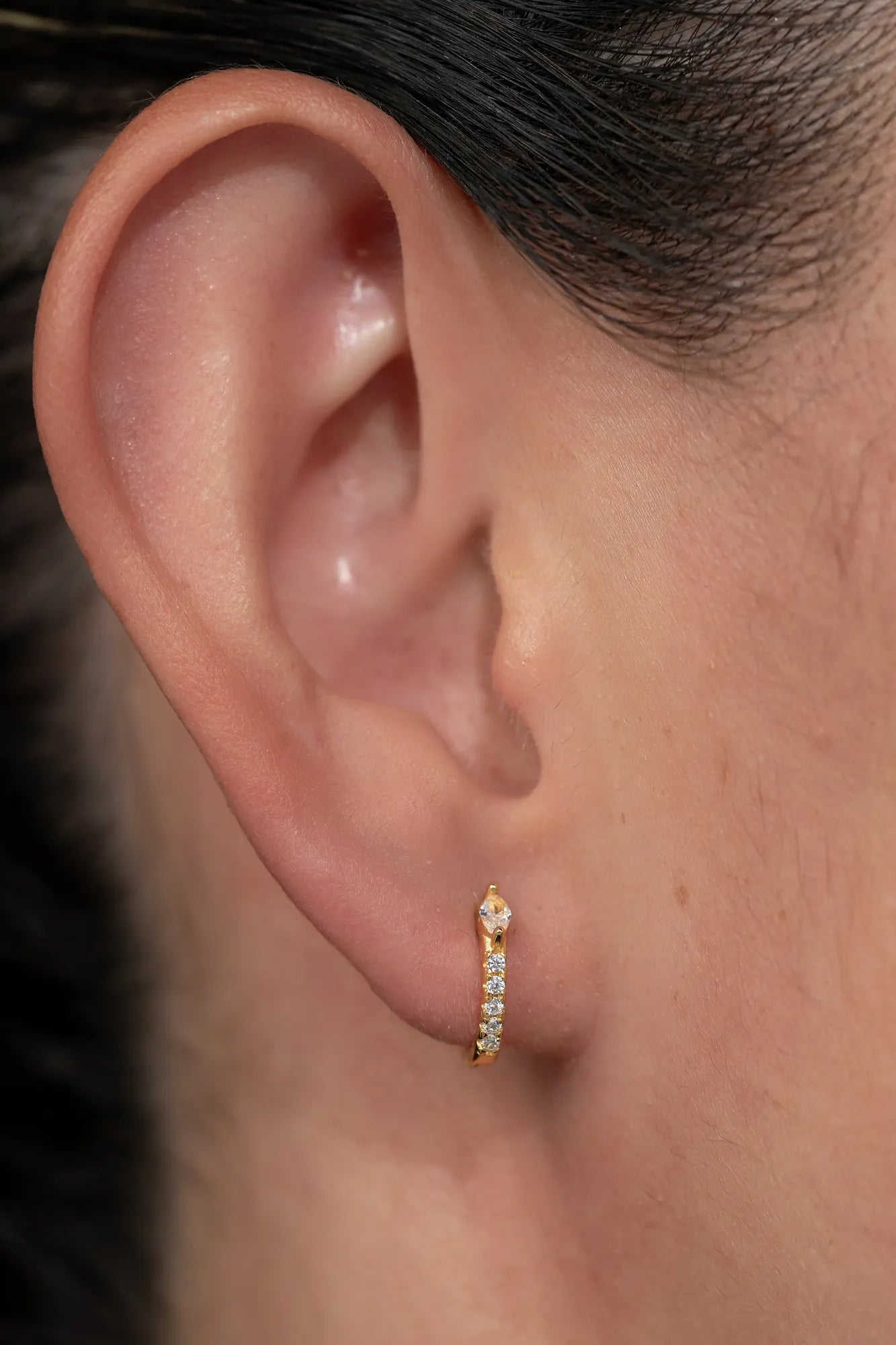 Model wearing petite gold huggie hoop earrings featuring a marquise-cut crystal accent and a row of sparkling stones along the front. 