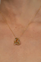 Close up of model wearing a delicate gold-tone necklace featuring an interlocking infinity knot pendant suspended from a fine chain. The minimalist design sits at the collarbone for an elegant, everyday look.