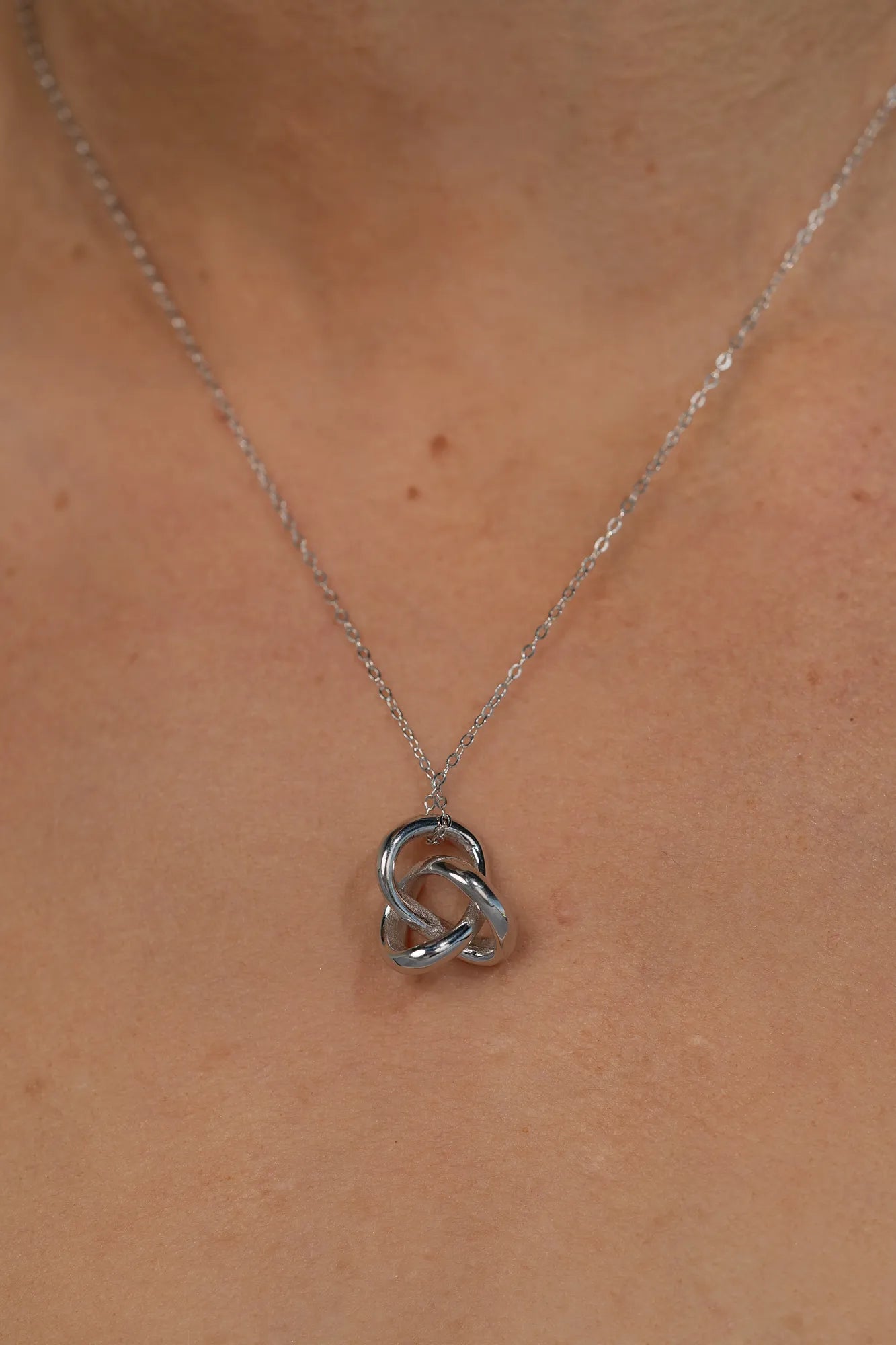 Close up of model wearing a delicate silver-tone necklace featuring an interlocking infinity knot pendant suspended from a fine chain. The minimalist design sits at the collarbone for an elegant, everyday look.