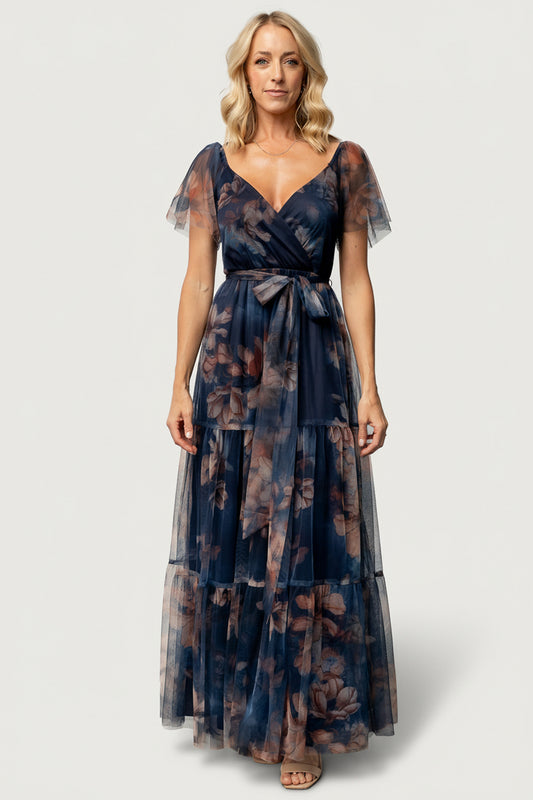 Front view of model wearing Nicola tulle maxi dress in dark blue floral print with surplice neckline and tiered skirt, flowing silhouette and raw hem.