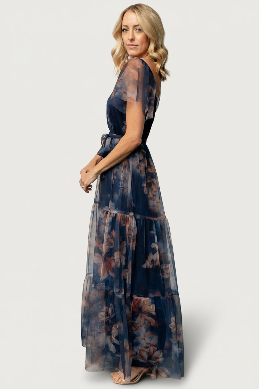 Studio image of Nicola tulle maxi dress in dark blue floral, styled with a subtle waist tie and soft tiered hem to demonstrate overall silhouette.