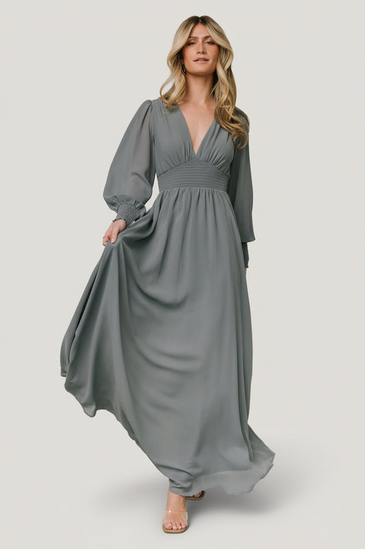 Model wearing Olivia Maxi Dress in dusty blue chiffon, front view showing deep V-neck, smocked waist, and flowing maxi skirt with soft movement.