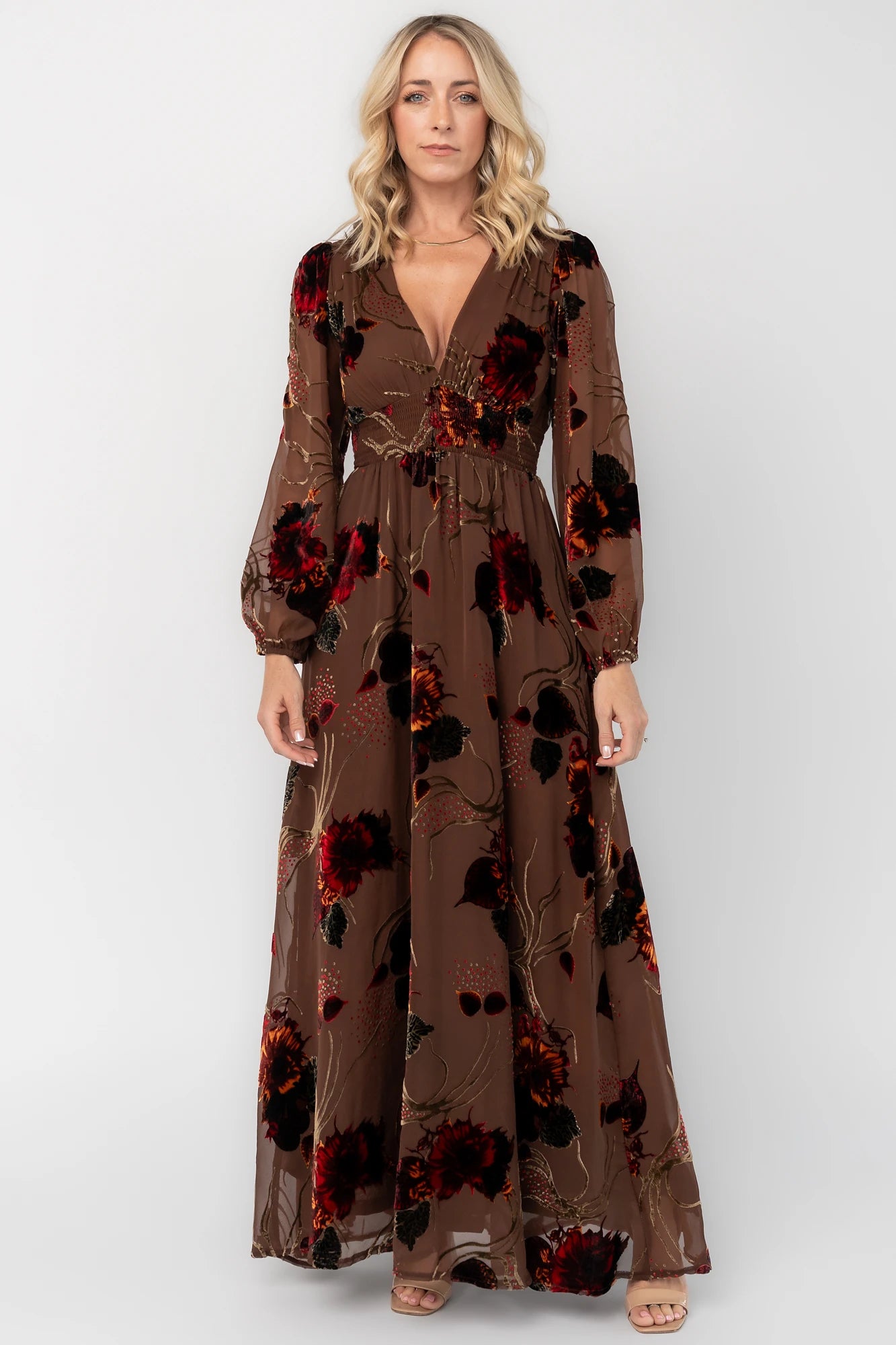 Brown burnout velvet maxi dress with a deep V-neckline, long sleeves, smocked waistband, and flowing asymmetrical hem with gold and burgundy floral accents.