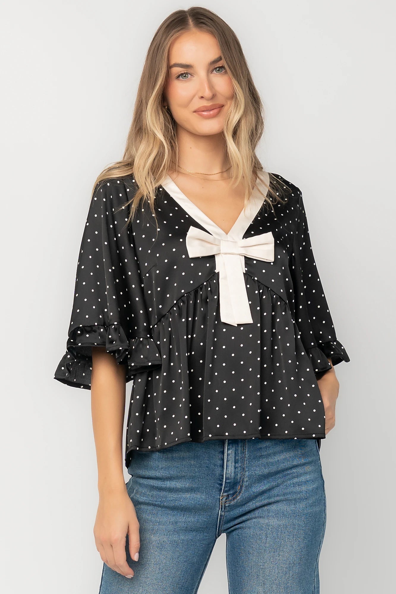 Black satin polka dot peplum top with an ivory contrast collar, decorative bow, V-neckline, and elbow-length ruffle sleeves.