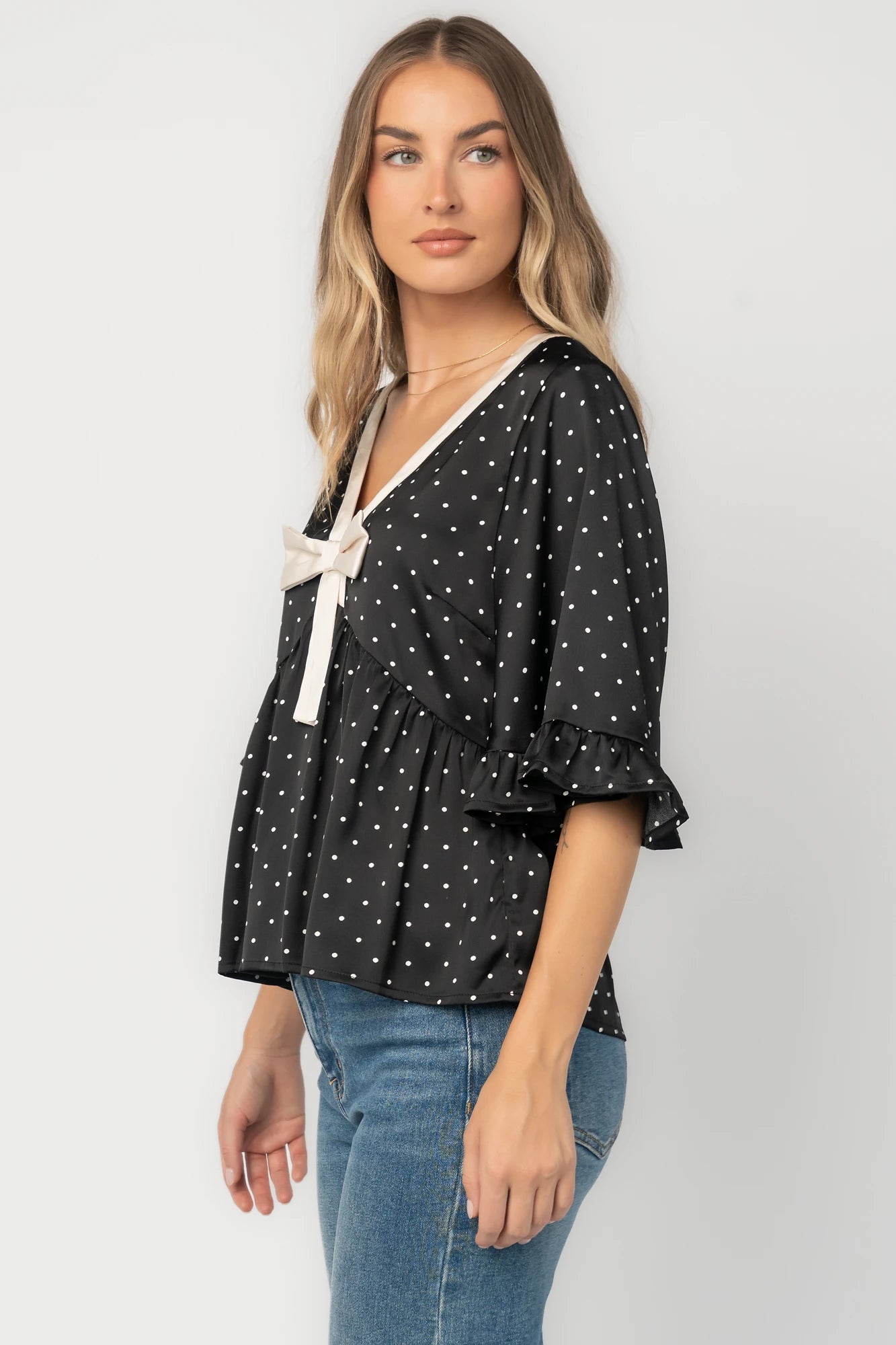 Black satin polka dot peplum top with an ivory contrast collar, decorative bow, V-neckline, and elbow-length ruffle sleeves.
