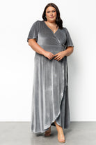 Full-length side profile of the dusty blue velvet wrap dress highlighting wrap tie at waist, tulip hem, and gentle movement of the skirt fabric in soft lighting.