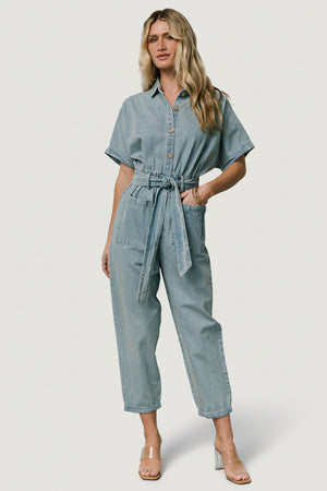 Back view of Mateo jumpsuit showing back patch pockets, tapered cuffed pant leg, and washed denim texture on model.