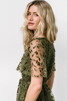 Side view of the olive dress highlighting the left-side slit, short unlined sleeve, and the embossed vine pattern flowing along the skirt.
