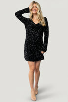 Front view of model wearing Mira black velvet sequin short dress with V-neck, long sleeves, and short hem, studio lighting.