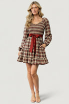 Front view of Stina Plaid Mini Dress on model showing V-neck smocked bodice, elastic cuffs, two-tiered short skirt, and waist sash.