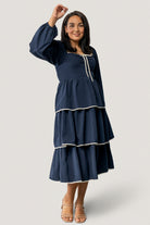 Front view of model wearing navy Barcelona tiered midi dress with sweetheart neckline, decorative bow, and ivory contrast trim at hem and sleeves.