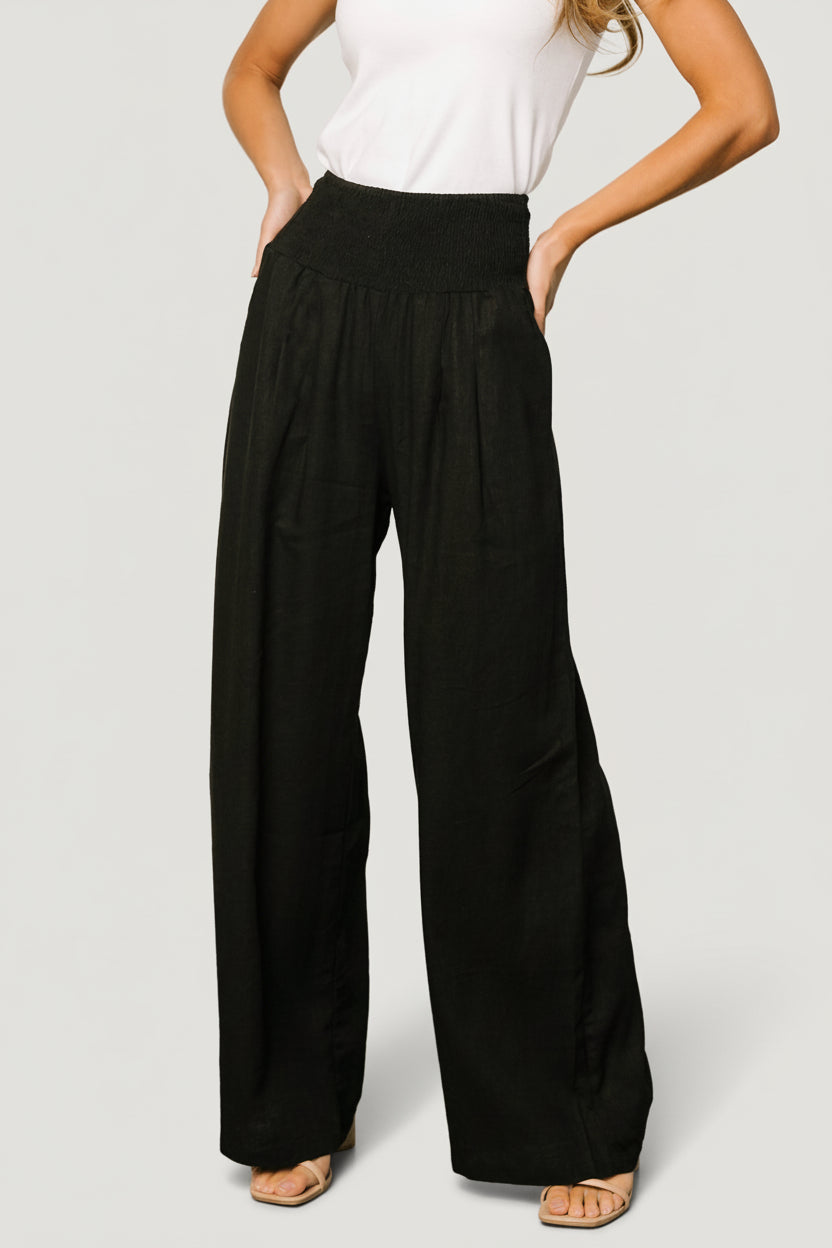 Front view of Parvati smocked high-waist wide-leg pants in black, showing smocked waistband, side pocket and fabric drape.