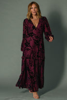 Model wearing mulberry velvet burnout embossed maxi dress, long sleeves, plunging V-neck, ruffle tiered skirt flowing to ankles.