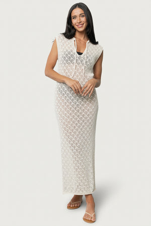 Model wearing the Calypso natural crochet cover-up standing on sand, showing scoop V-neck with tie and relaxed straight fit, unlined.