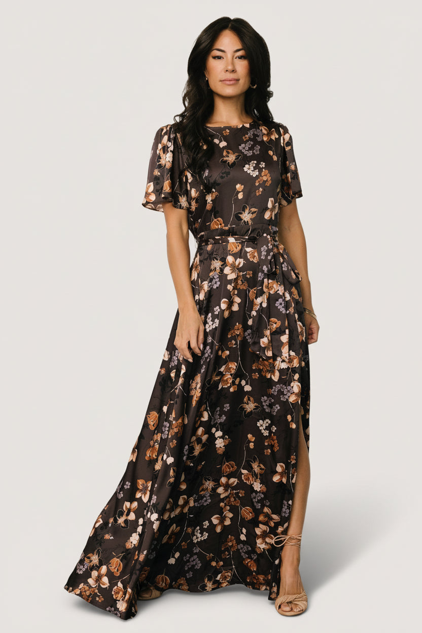 Front view of brown floral satin maxi dress on mannequin featuring rounded neckline, short flutter sleeves, removable belt tie, and left side slit.