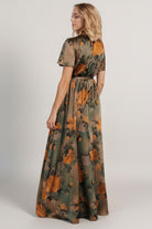 Side angle of olive floral satin maxi dress showcasing the left-leg slit and fluid movement of the skirt while walking.