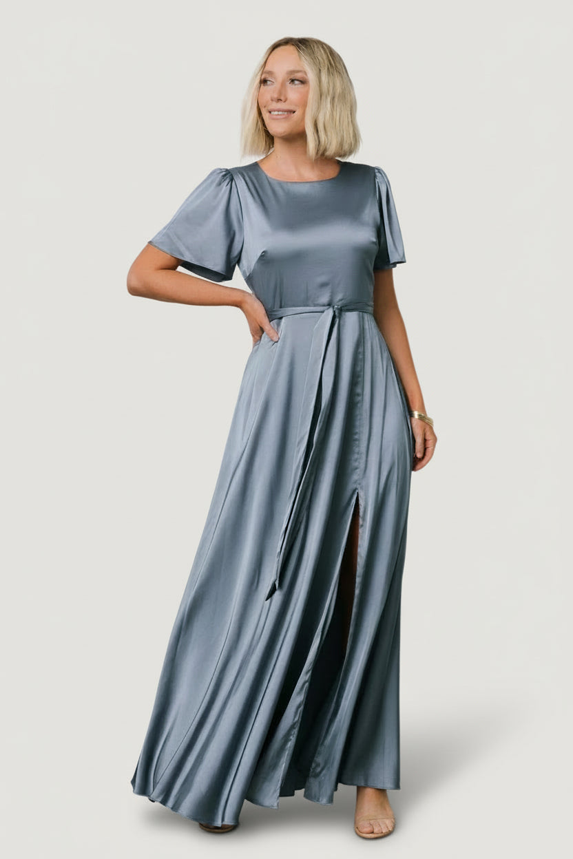 Model wearing Agnes Satin Maxi Dress in whisper blue, front view showing rounded neckline, short flutter sleeves, removable waist tie, and left slit.