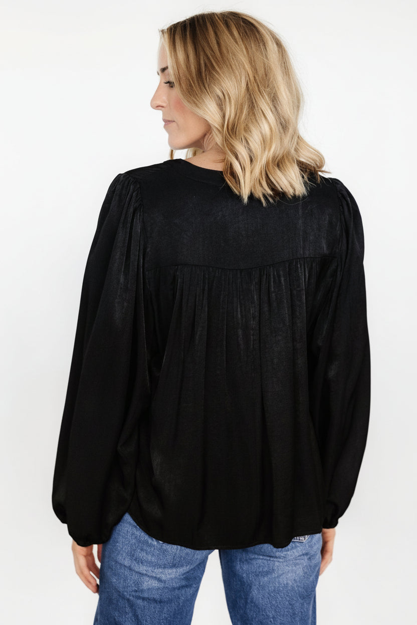 Styled outfit image with the black Alina blouse paired with fitted trousers, illustrating how the relaxed blouse tucks neatly for a polished look.
