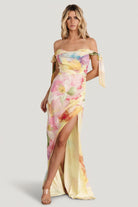 Front view of yellow multi-floral off-shoulder chiffon maxi dress showing cowl neckline, adjustable bow straps, and tulip skirt in motion.