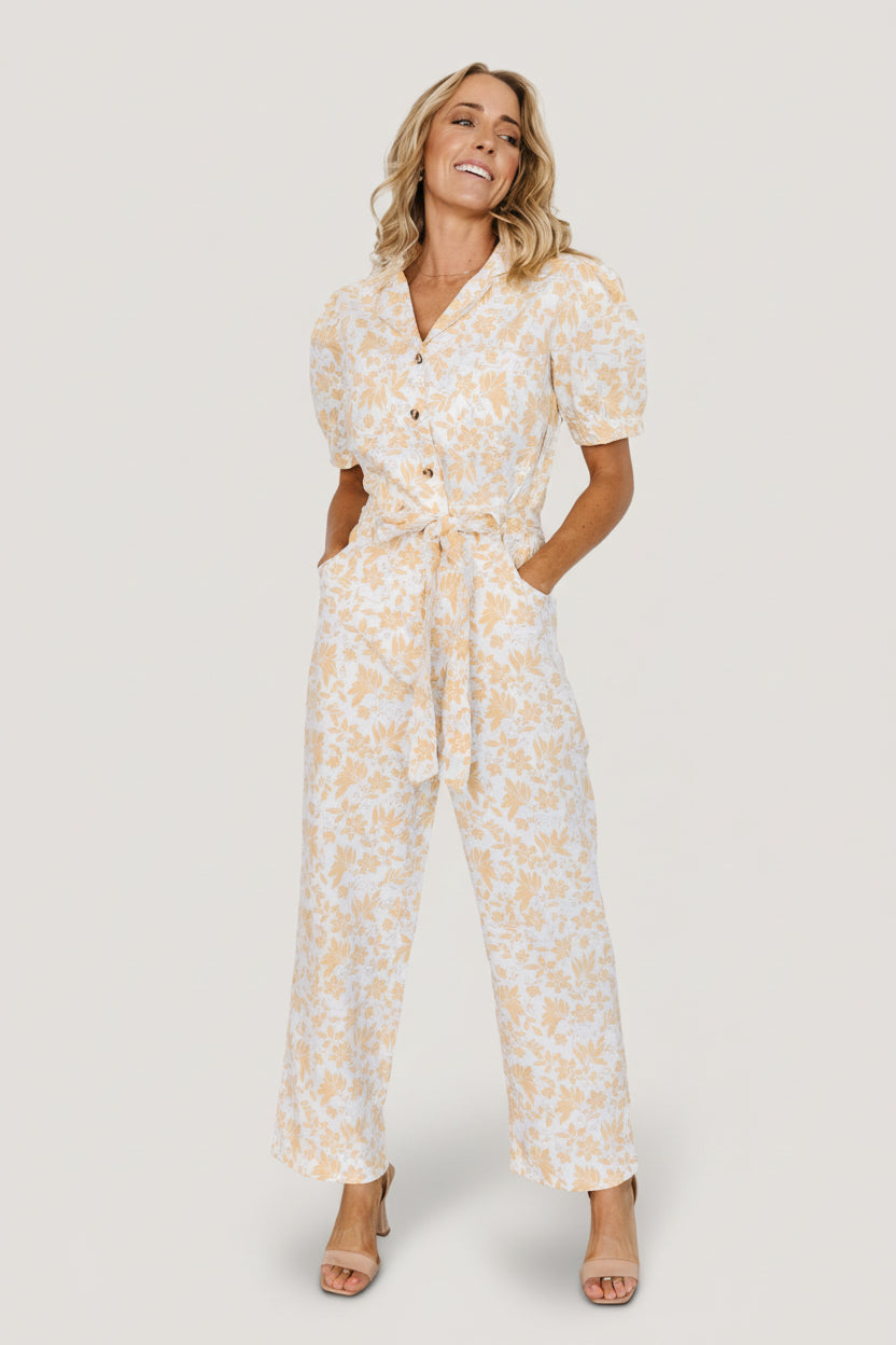 Front view of Annalise belted utility jumpsuit in white with muted yellow floral print, button-front and puff sleeves (model shot).