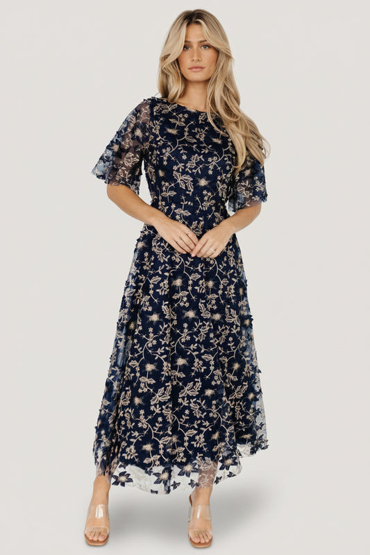 Front view of navy tulle maxi dress with gold embroidered floral pattern, round neckline, short sheer sleeves and flowing full-length skirt.