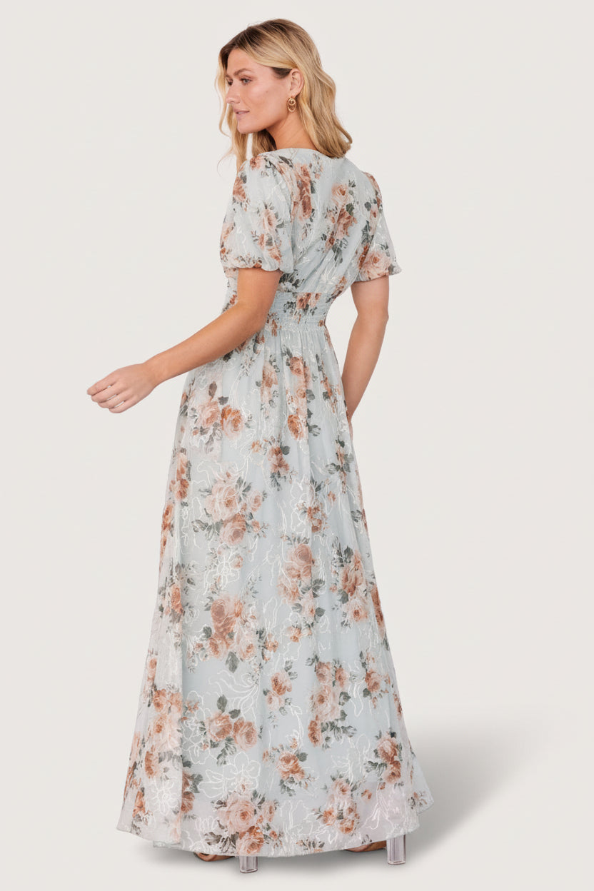 Model wearing sage floral maxi dress in motion, showing natural skirt drape, V-neck silhouette, short puff sleeves, and embroidered accents.