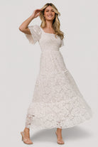 Front view of off-white lace maxi dress with smocked bodice, ruffled square neckline, short bell sleeves, and tiered skirt, studio lighting.