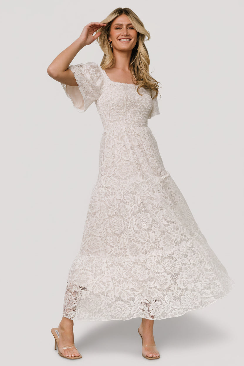 Front view of off-white lace maxi dress with smocked bodice, ruffled square neckline, short bell sleeves, and tiered skirt, studio lighting.