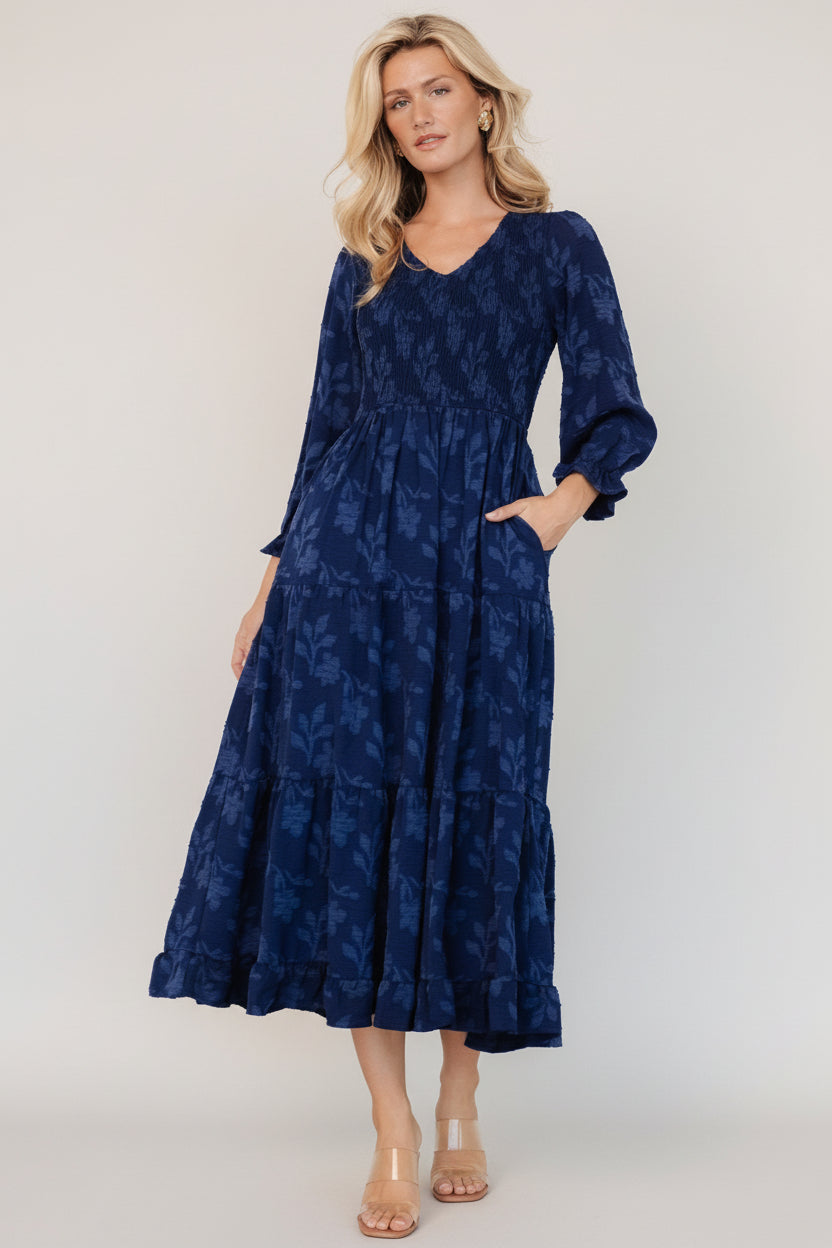 Front view of navy floral smocked midi dress showing V-neck, elastic smocked bodice, tiered skirt, and side seam pocket details.