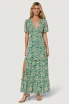Front view of model wearing green floral chiffon deep V maxi dress with tiered ruffle skirt and right-side slit, shown full length.