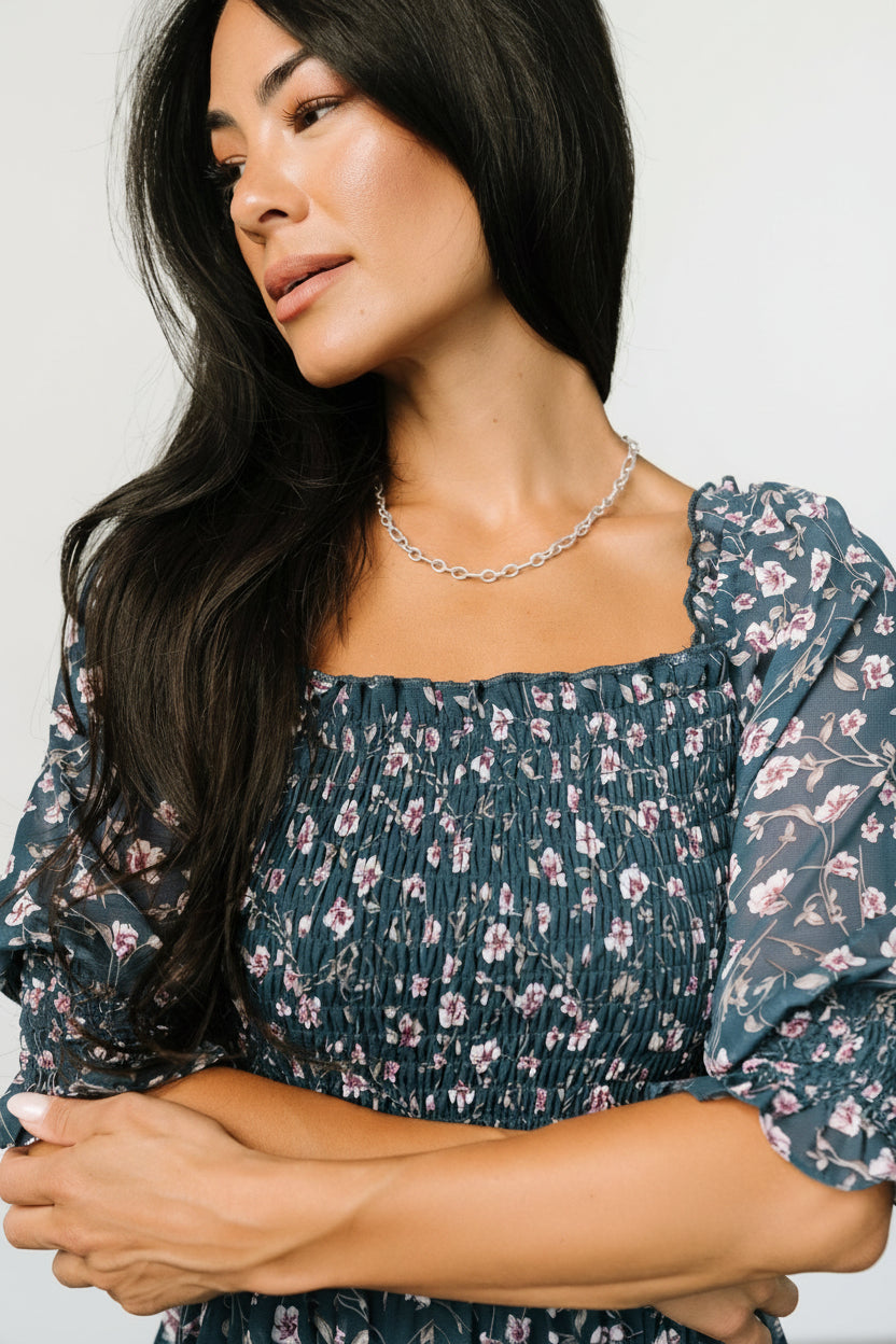 Model wearing Azlyn smocked short dress in blue multi floral, displaying tiered short skirt, detached waist tie with tassels, and semi-sheer sleeves.