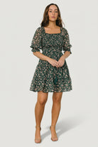 Front view of green and pink floral smocked short dress on a model, showing smocked bodice, ruffled square neckline and tiered short skirt for movement.